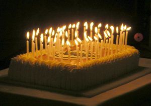 Neliön muotoinen vaalea syntymäpäiväkakku, jossa keskellä on Hengityslaitepotilaat ry:n vihreä logo. Kakussa kynttilöitä.