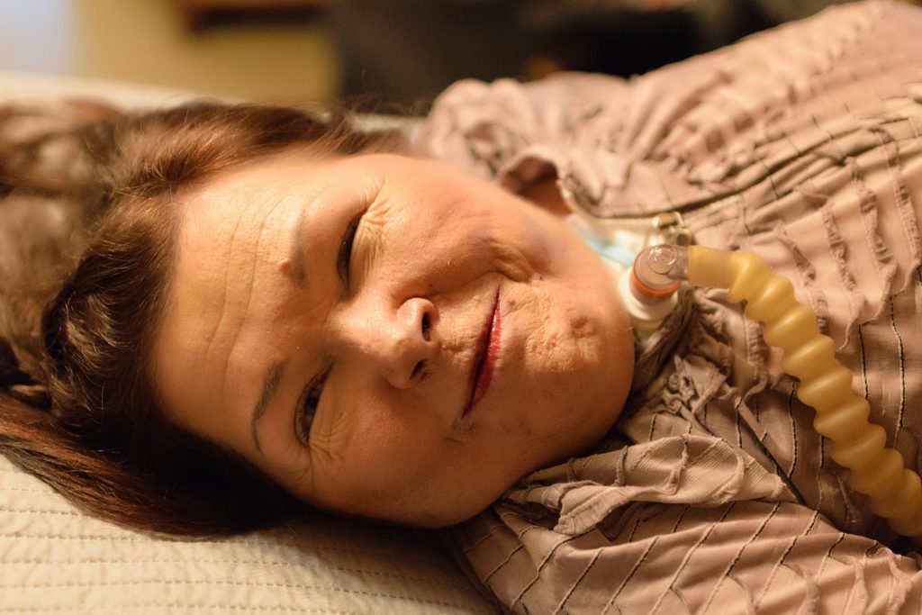 Hengityslaitetta käyttävä ruskeatukkainen nainen makaa sängyllä ja hymyilee kameralle