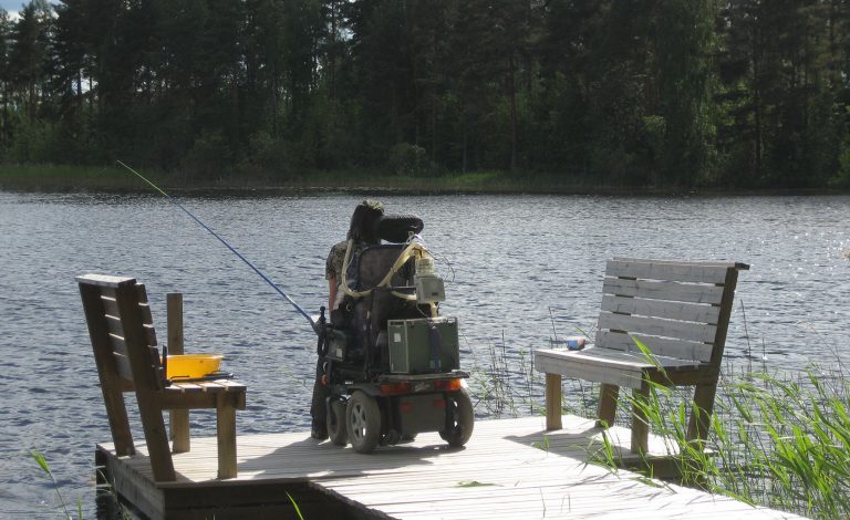 Hengityslaitetta käyttävä mies sähköpyörätuolissa kalastamassa laiturilla