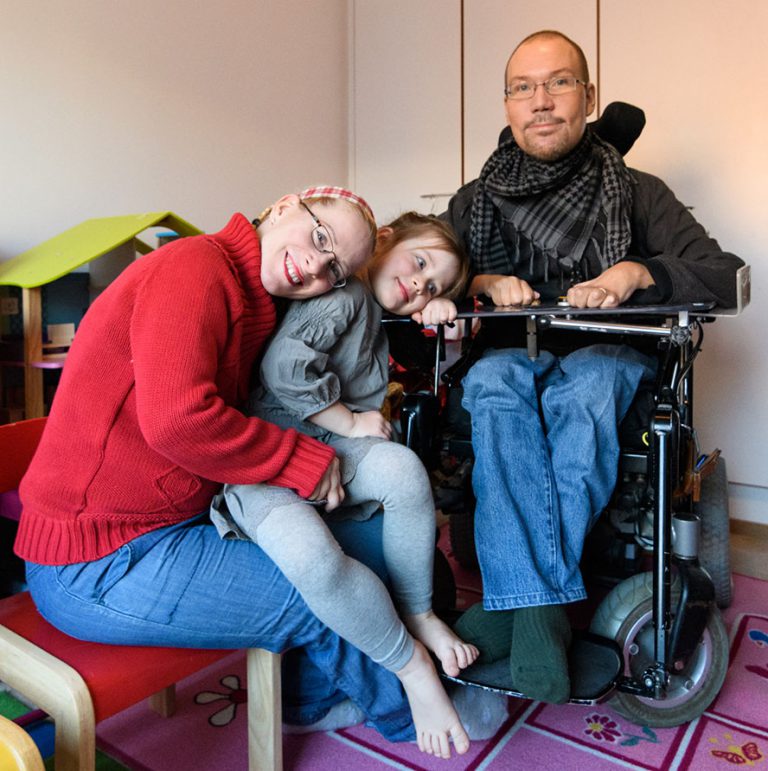Duchennea sairastava mies pyörätuolissa hengityskoneen kanssa, vierellään puoliso sekä lapsi, jotka ovat taivuttaneet päänsä kohti miestä