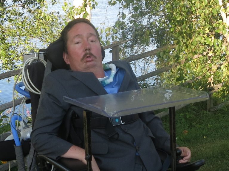 Jukka Sariola harmaassa puvussa pyörätuolissa koivikon keskellä