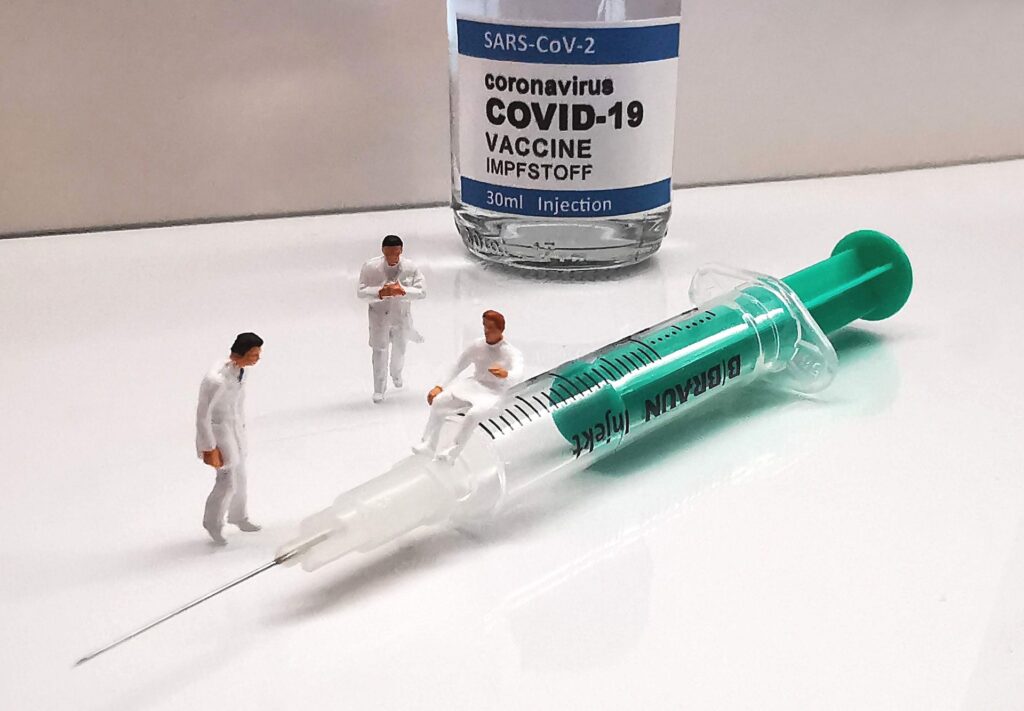 pöydällä covid19-rokotepurkki sekä neula, jonka ympärillä on terveydenhoidon asuihin puettuja pieniä muovinukkeja kolme kappaletta