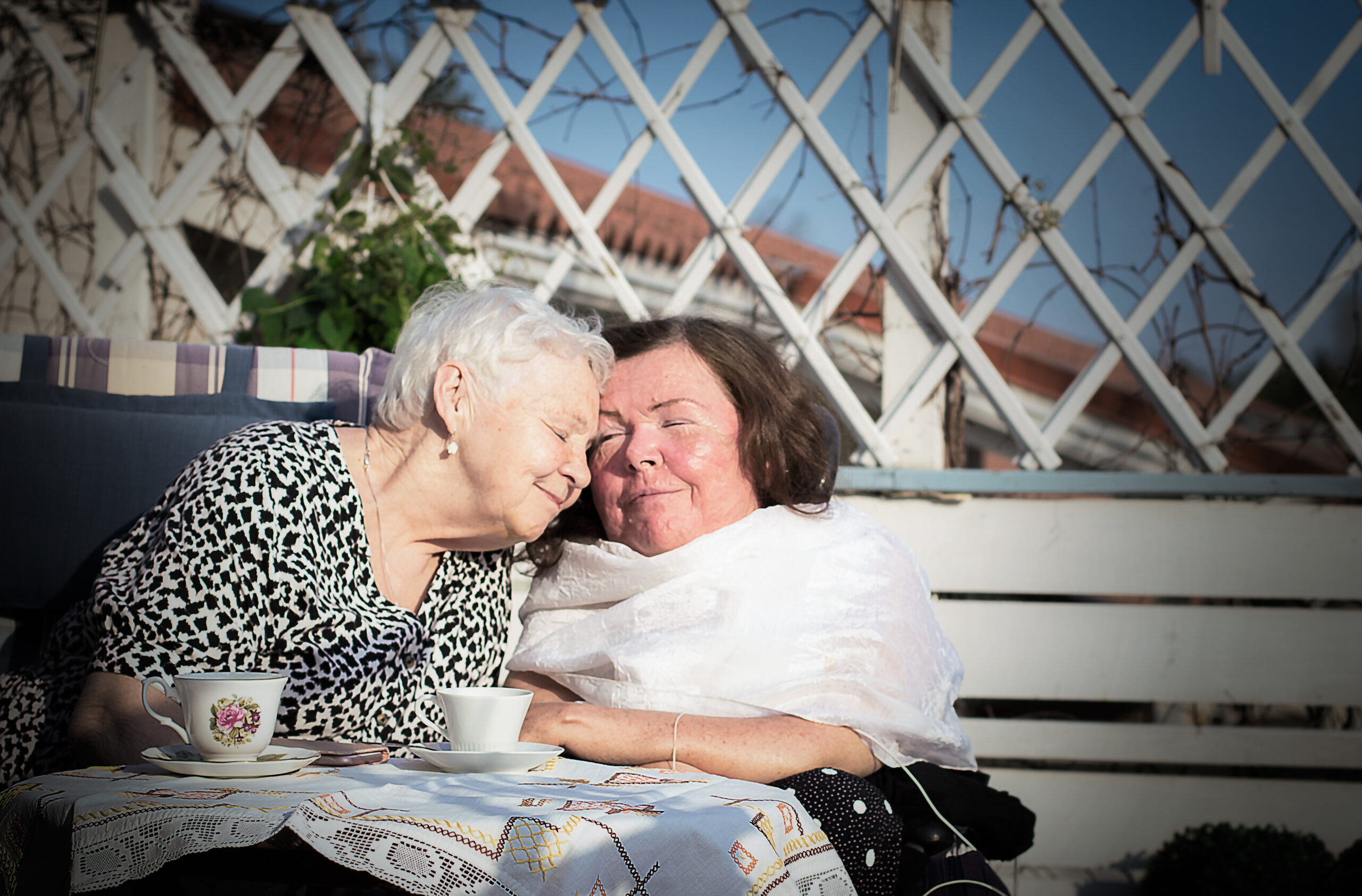 Kaksi eläkeikäistä naista puutarhapenkillä välittämistä ilmaisevassa asetelmassa toisiinsa nähden, hymy huulilla