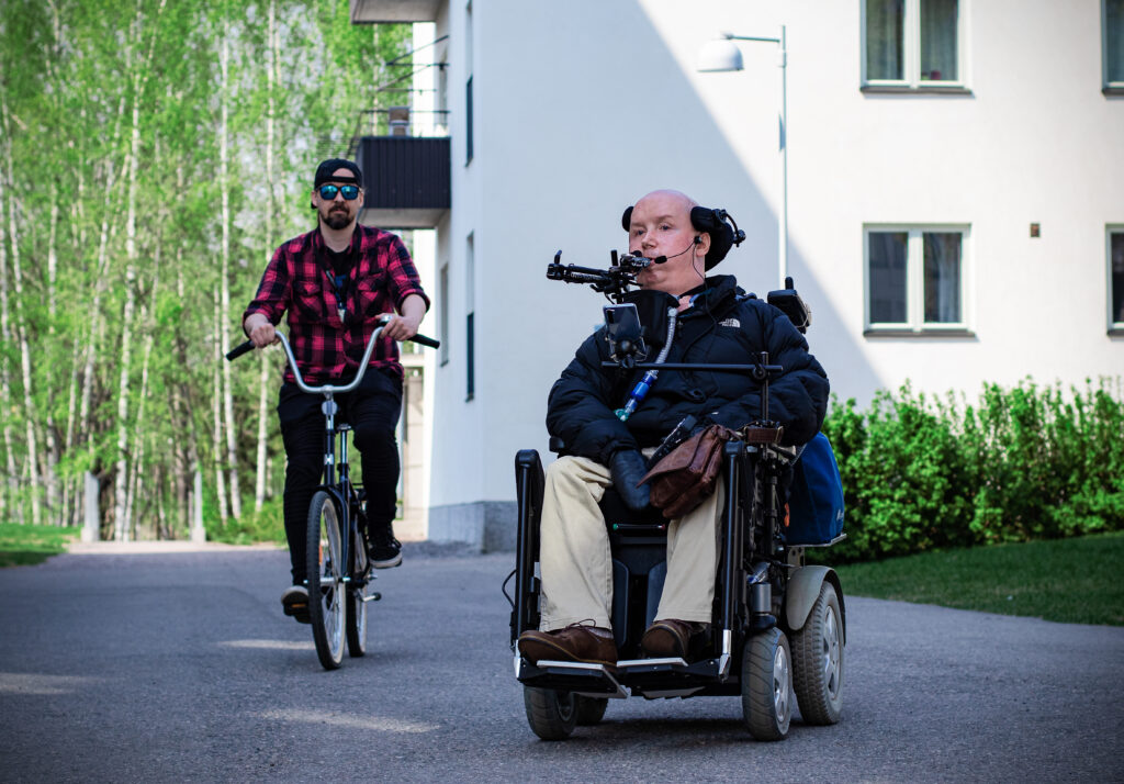 sähköpyörätuolilla liikkuva mies, jonka vierellä polkupyörällä ajava mies kesällä kerrostalon pihatiellä