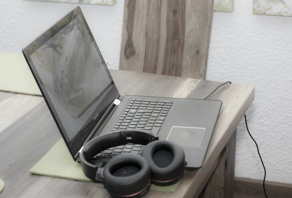 Kannettava tietokone ja kuulokkeet puisella harmahtavalla pöydällä. Taustalla näkyy samaa puuta olevaa selkänöjallinen tuoli
