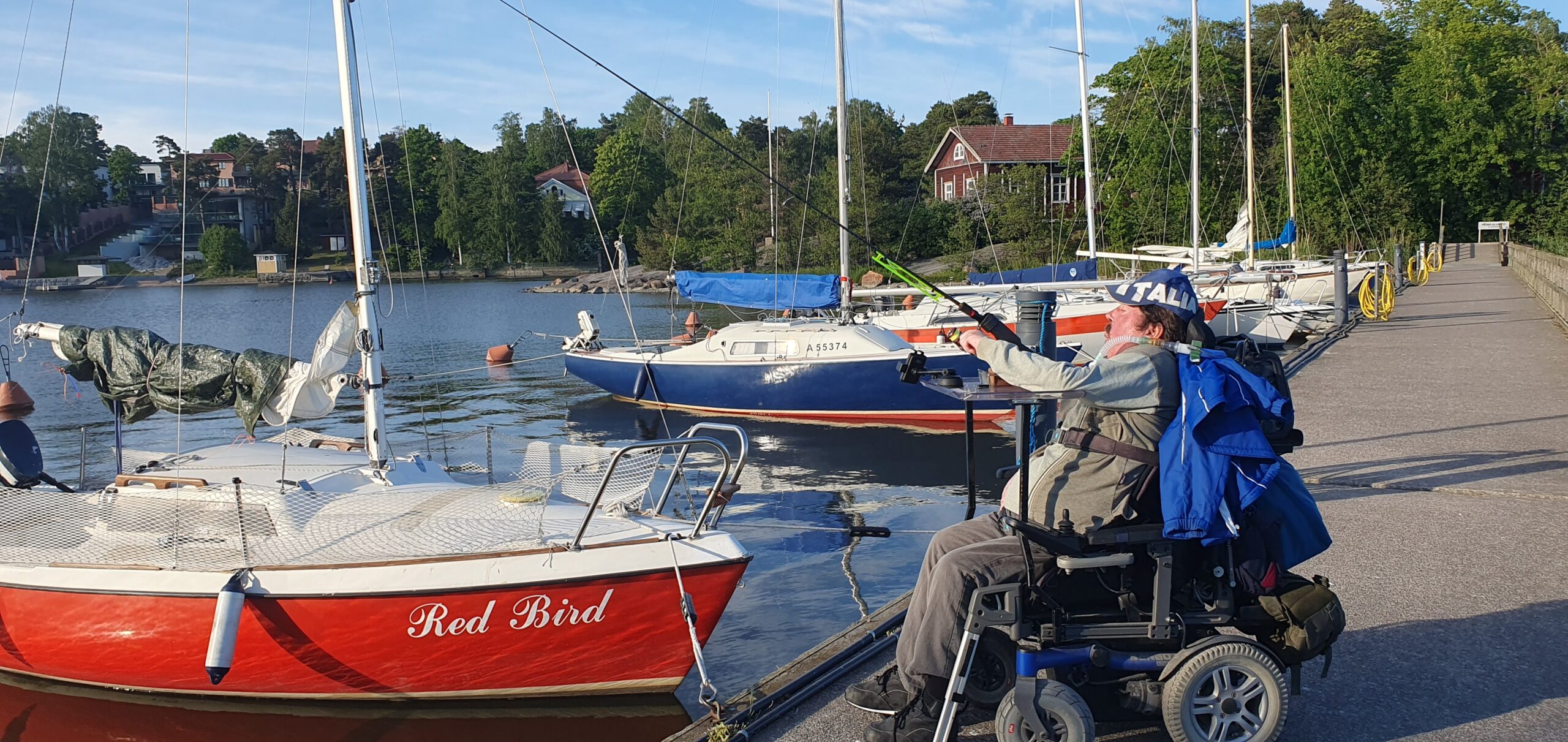 hengityshalvusstatuksen omaaja pyörätuolissa kalastamassa laiturilla virvelillä. Taustalla punainen purjevene ja muita purjeveneitä